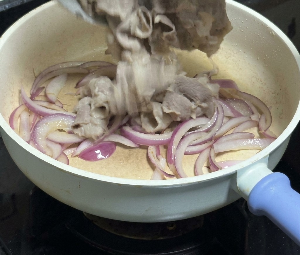 Thinly sliced beef being poured from a white bowl into a frying pan filled with translucent red onion strips.