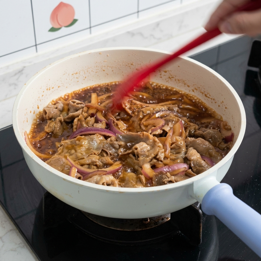 Thinly sliced beef and red onion rings being tossed with a red spatula in a white pan filled with bubbling brown teriyaki sauce.