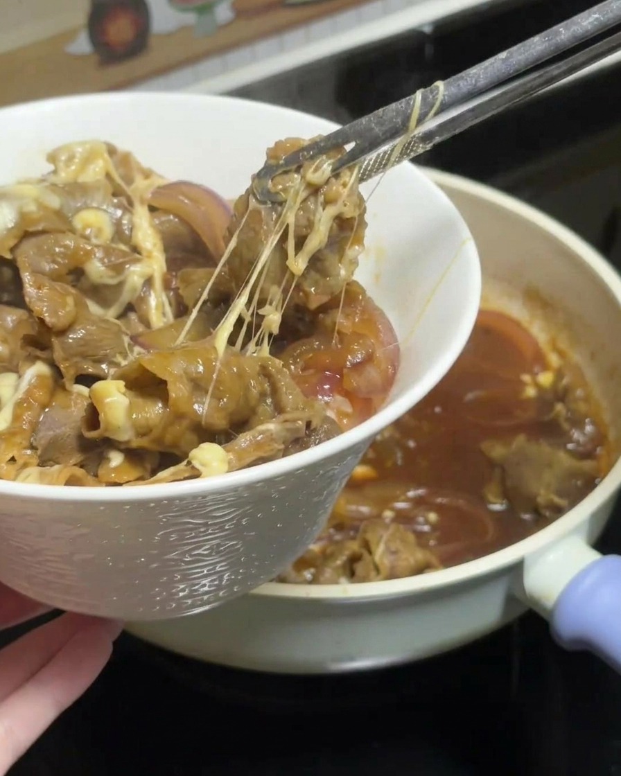 Tongs lifting a portion of cheesy teriyaki beef and onions from the pan, showing long cheese strings, and placing it onto a bowl of white rice.