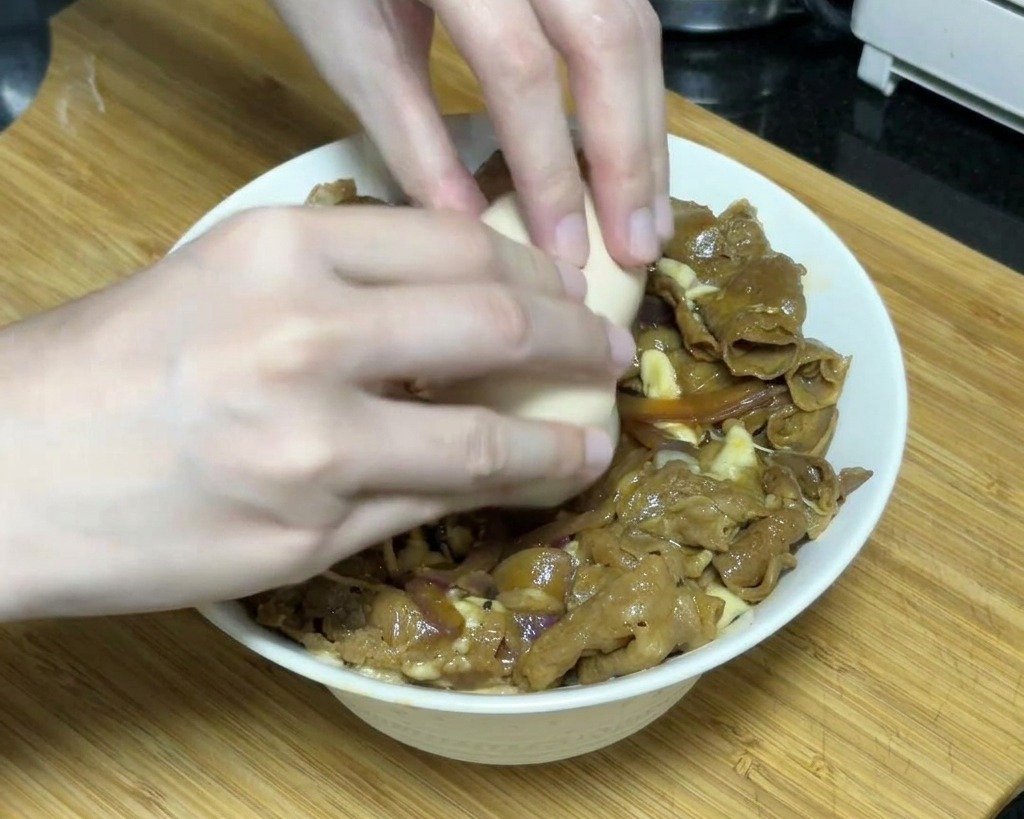 A pair of hands carefully cracking a soft-set onsen egg over a bowl of teriyaki beef topped with melted cheese.