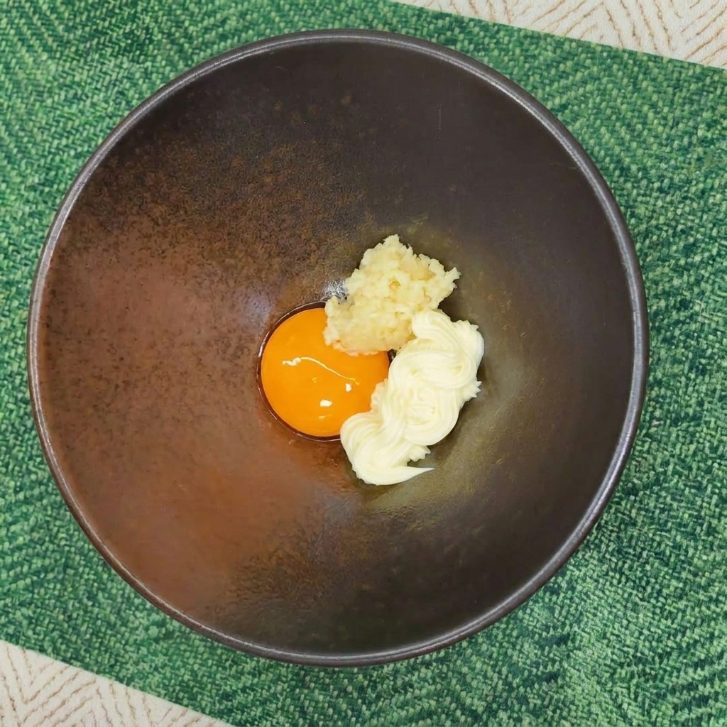 A dark ceramic bowl containing a raw egg yolk, a dollop of mayonnaise, and minced garlic on a green placemat.