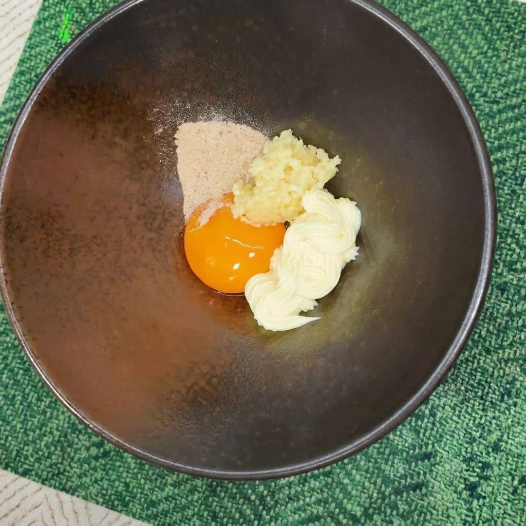 Instant noodle seasoning powder poured over an egg yolk, mayonnaise, and minced garlic in a dark ceramic bowl.