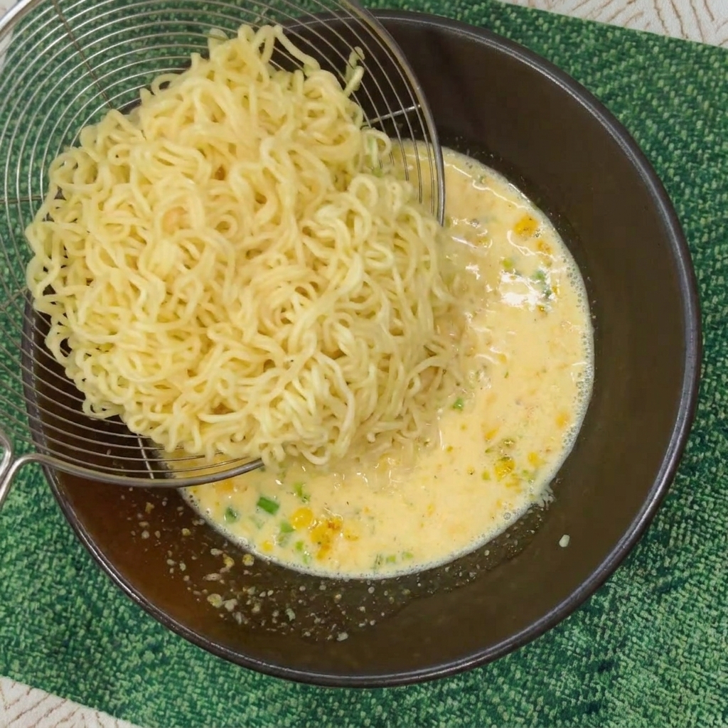 A metal strainer transferring cooked wavy instant noodles into a dark bowl filled with creamy, yellow broth.