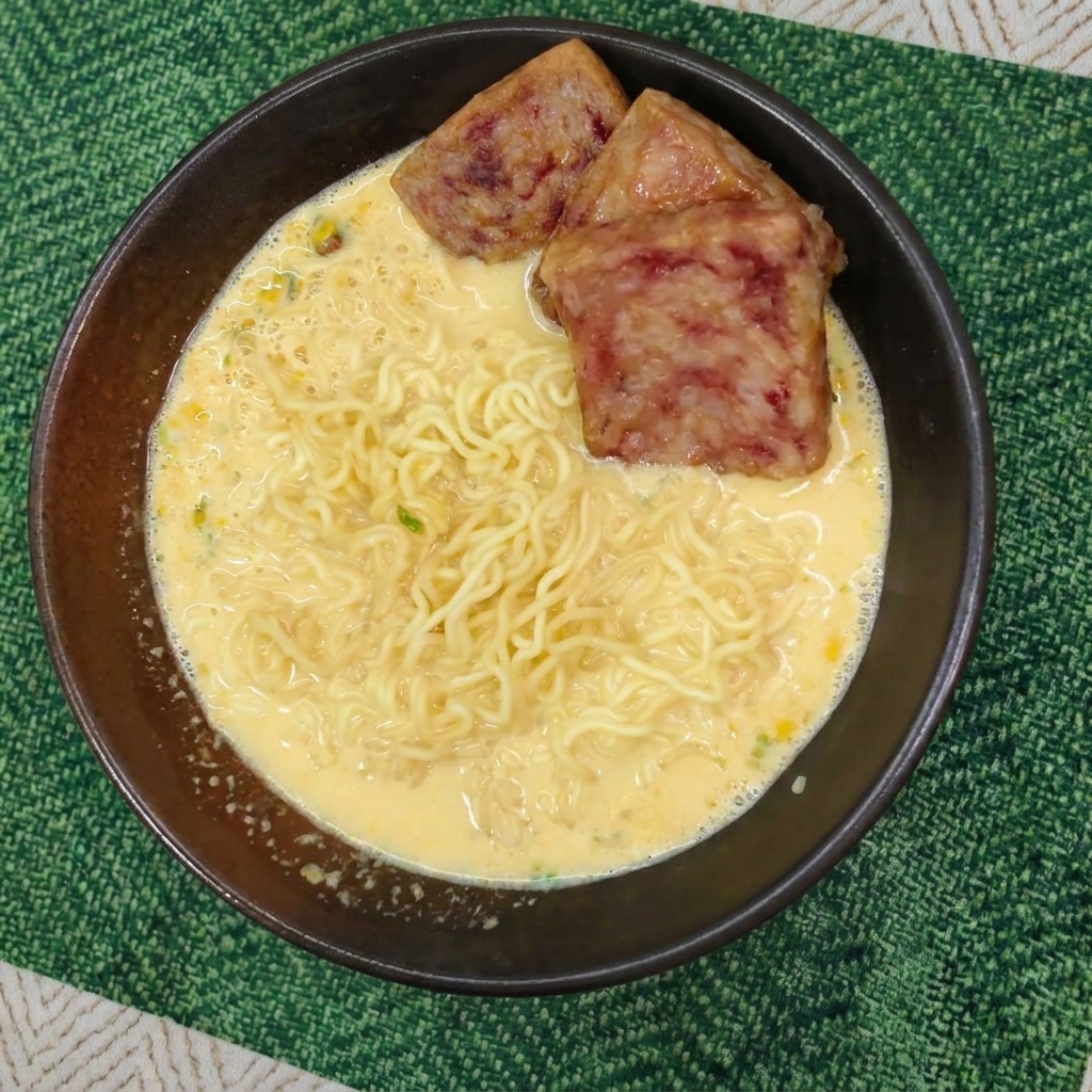 Cooked wavy noodles swimming in a creamy yellow broth, topped with three slices of browned spam in a dark bowl.