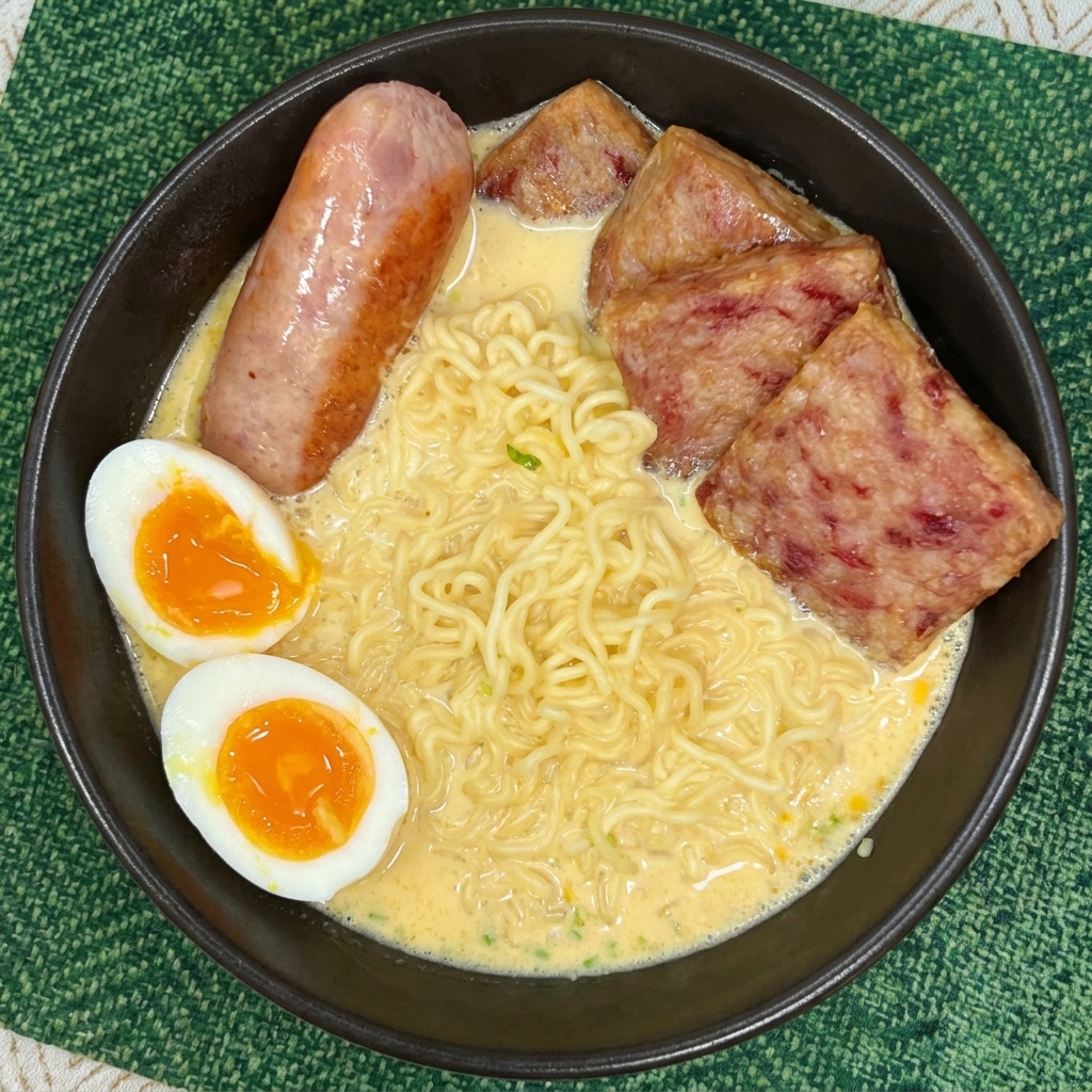 A bowl of creamy ramen noodles topped with slices of spam, a cooked sausage, and two soft-boiled egg halves with runny yolks.