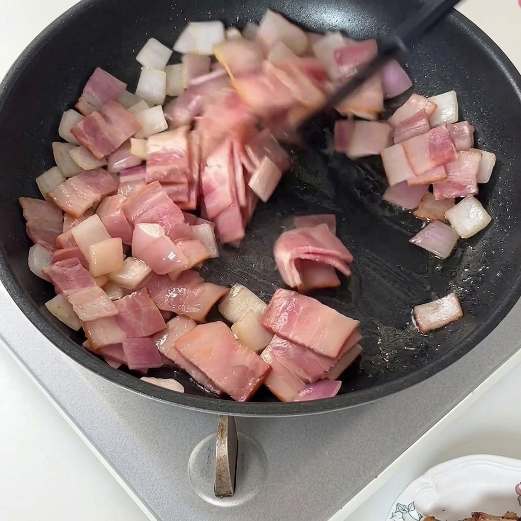 A spatula stirring cooked bacon pieces and onions in a frying pan.