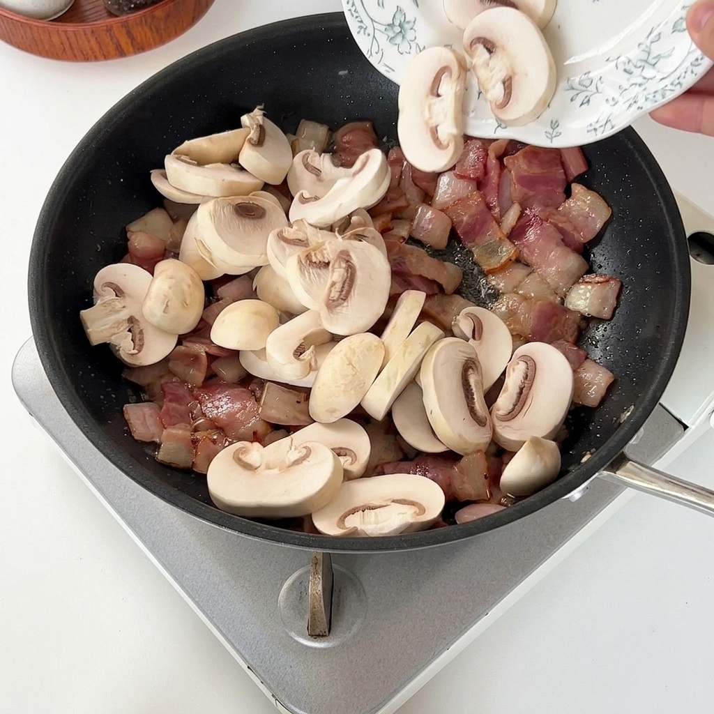Sliced white mushrooms being added from a plate into a hot pan with cooked bacon and onions.