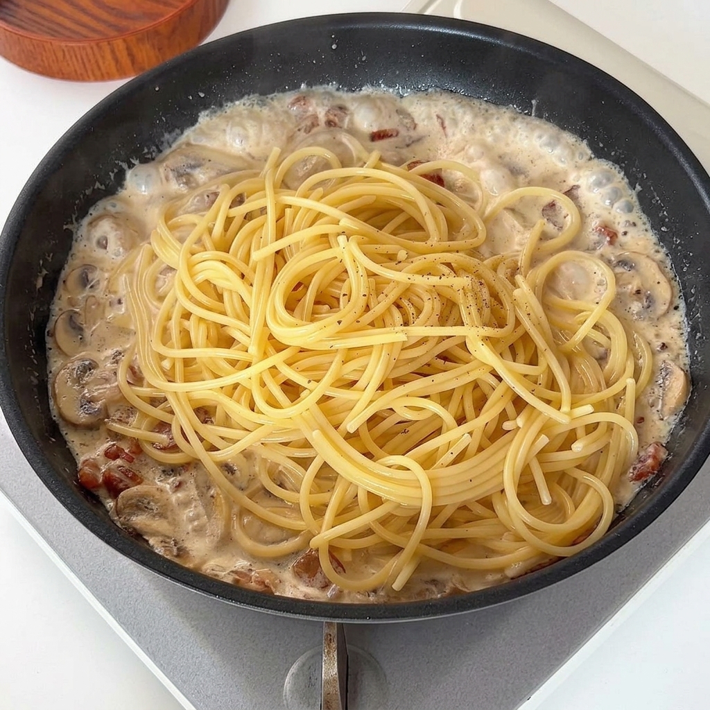 Cooked spaghetti noodles being placed on top of a creamy mushroom and bacon sauce in a frying pan, topped with black pepper.
