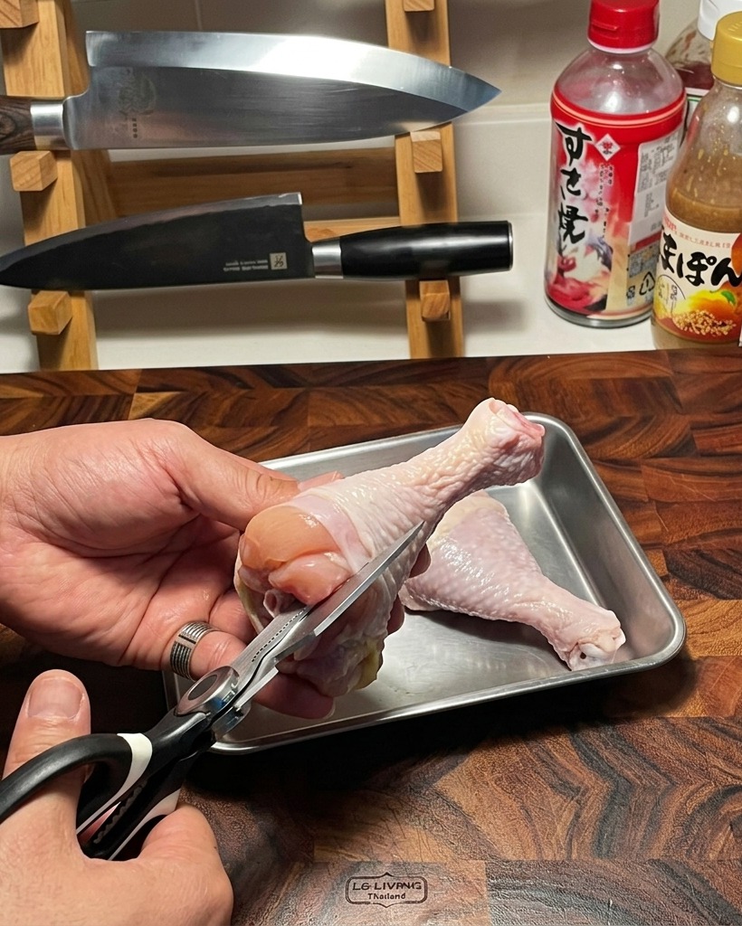 A pair of kitchen scissors being used to carefully extract the bone from a fresh, raw chicken thigh on a wooden cutting board.