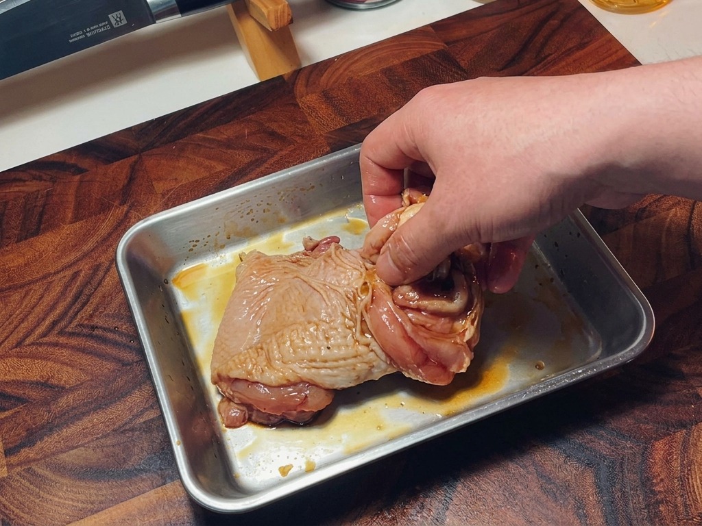 A hand mixing seasoned raw chicken thighs in a tray to ensure even distribution of the marinade.