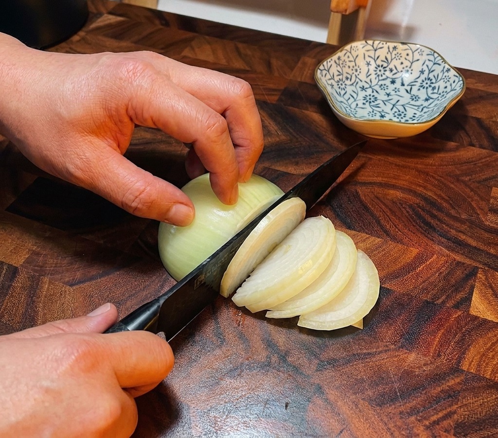 A chefs knife slicing through a white onion on a dark wooden cutting board.