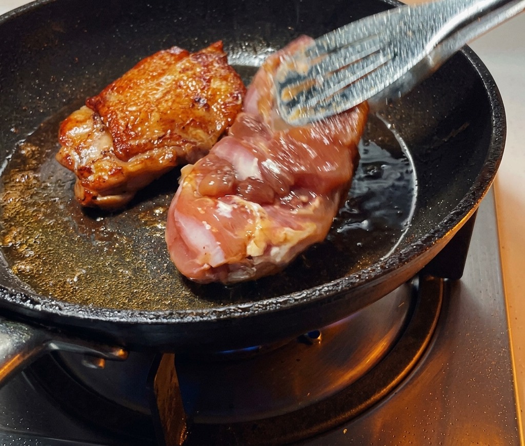 Tongs flipping a seared chicken thigh in a pan, revealing a perfectly browned and crispy skin.