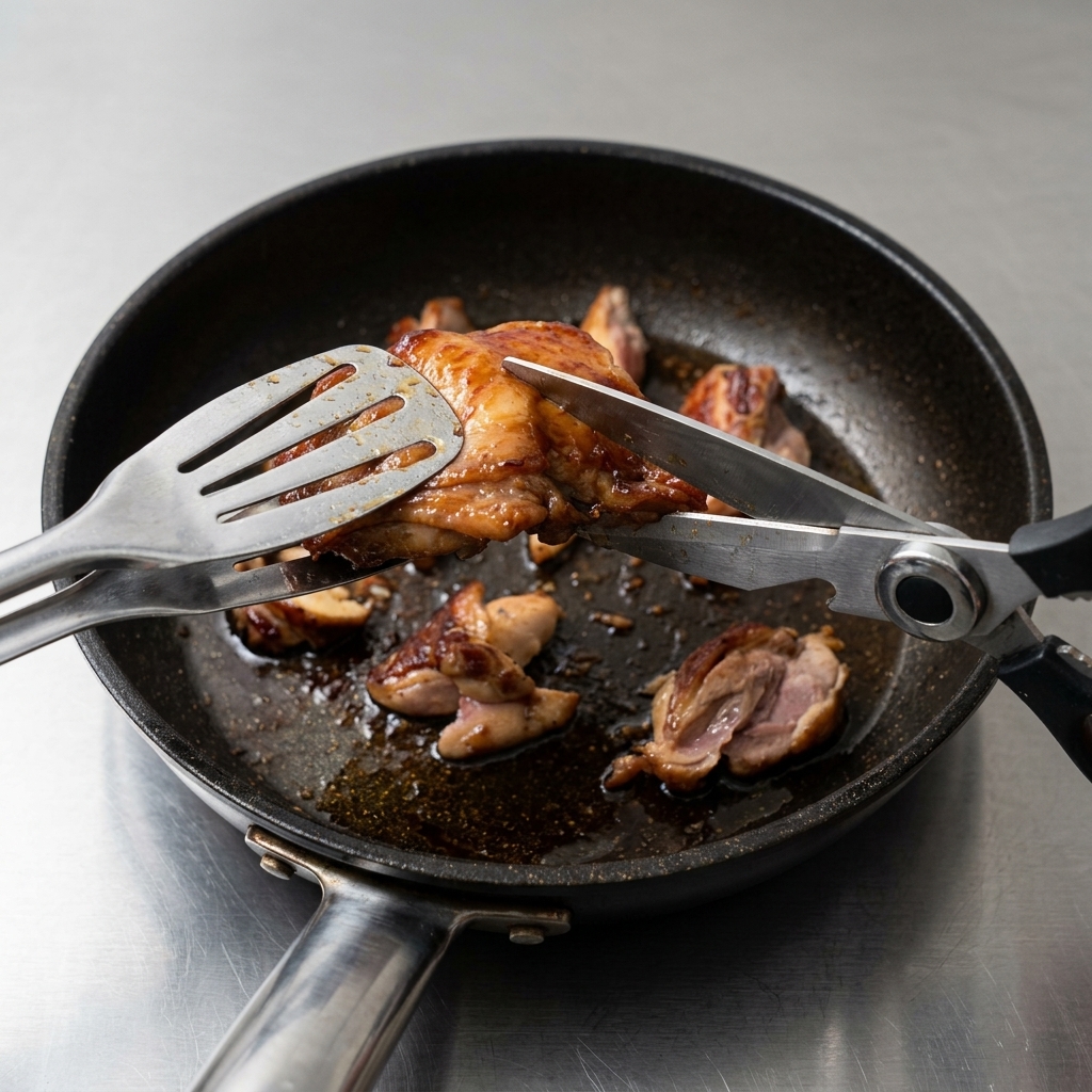 Kitchen scissors cutting through seared chicken thighs in a pan to create small, uniform morsels.