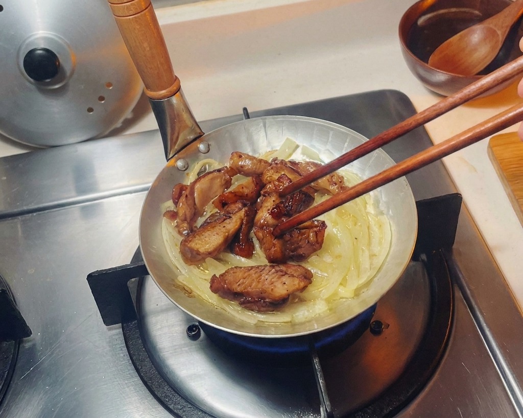 Seared chicken pieces being arranged with chopsticks over a bed of translucent onions in a small frying pan.