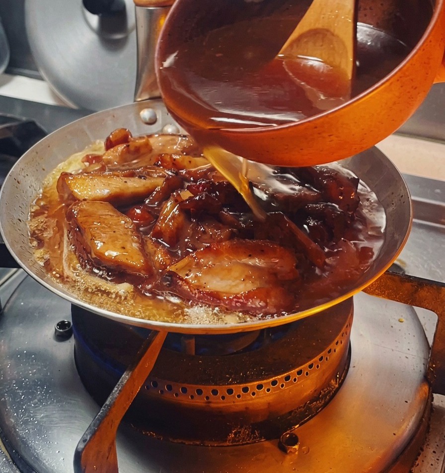 A dark savory sauce being poured from a small orange bowl into a pan filled with browned chicken pieces and onions.
