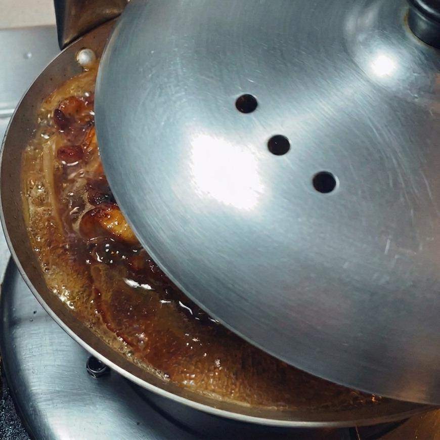 A polished metal lid being placed onto a shallow pan to trap steam and simmer the contents.