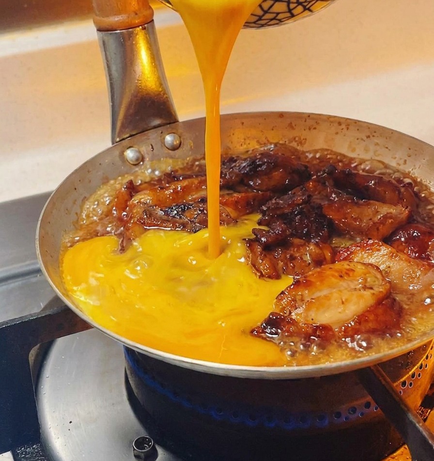 Golden-yellow beaten egg liquid being poured into a pan over simmering chicken pieces.