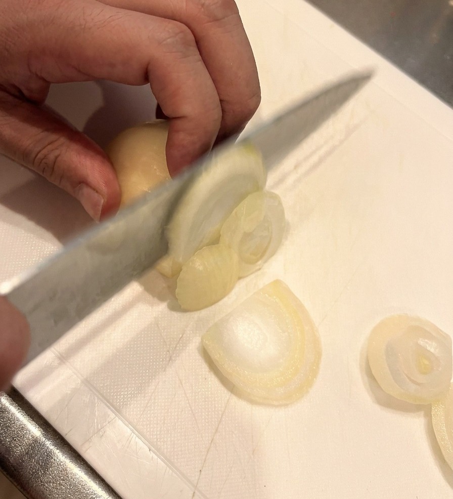 A sharp knife slicing a white onion into thin segments on a white cutting board, with a hand holding the onion securely.