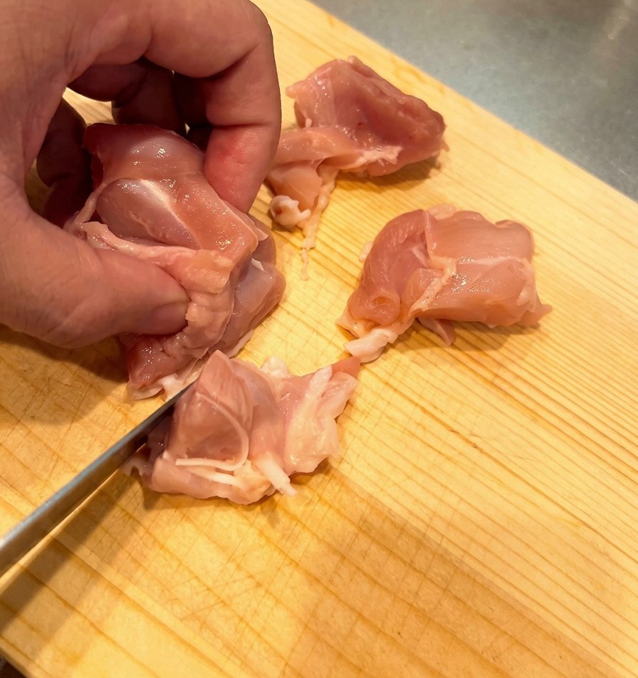 Raw chicken thigh meat being diced into small, uniform chunks on a light-colored wooden board.