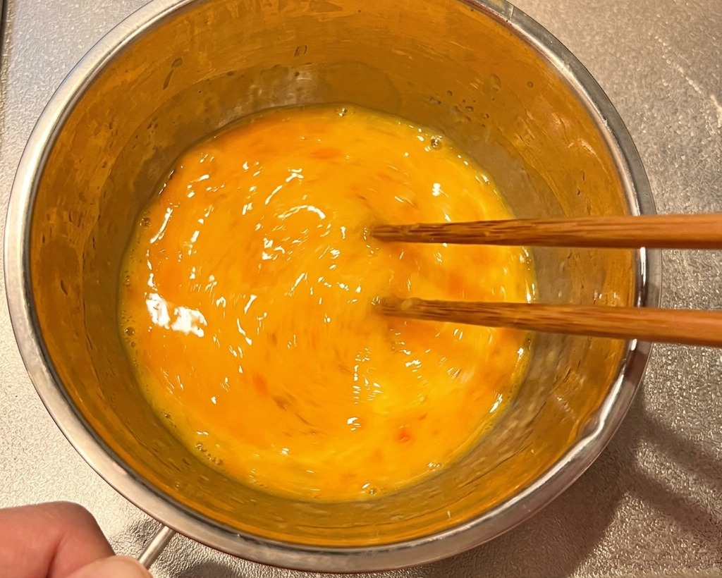 Whisked eggs in a stainless steel bowl being stirred with wooden chopsticks.