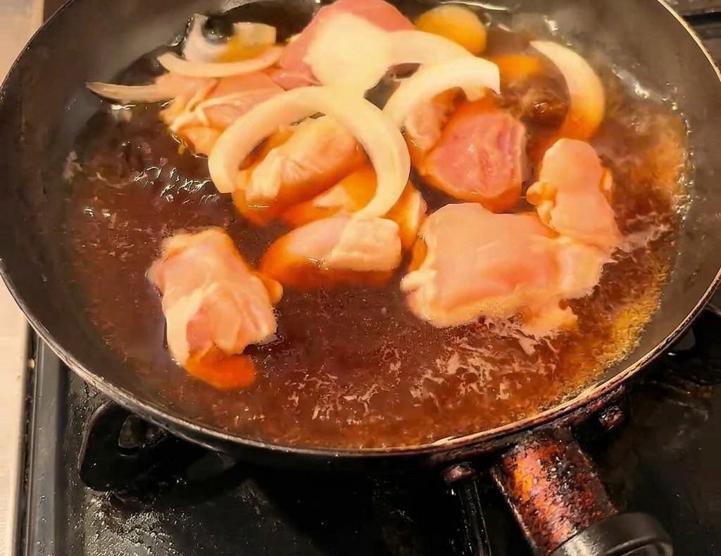 Chicken pieces and sliced onions simmering in a dark soy-sauce broth in a black frying pan.