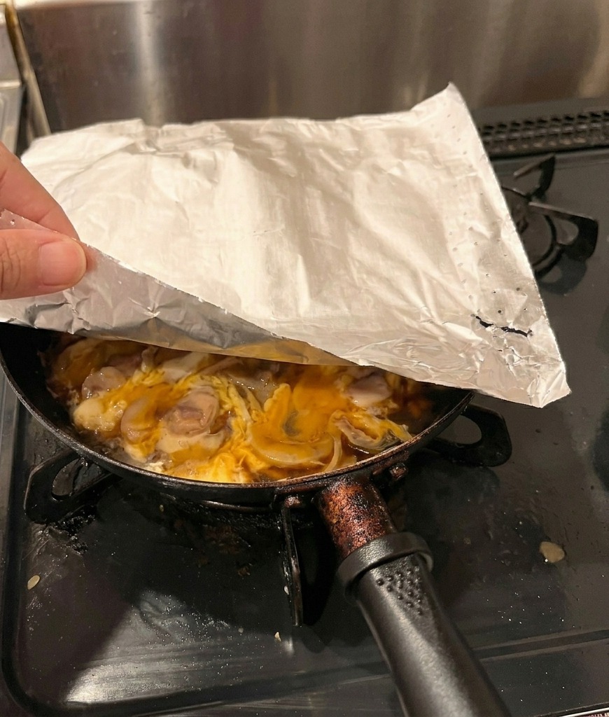 A hand placing a sheet of white parchment paper over a pan of chicken and partially cooked eggs.