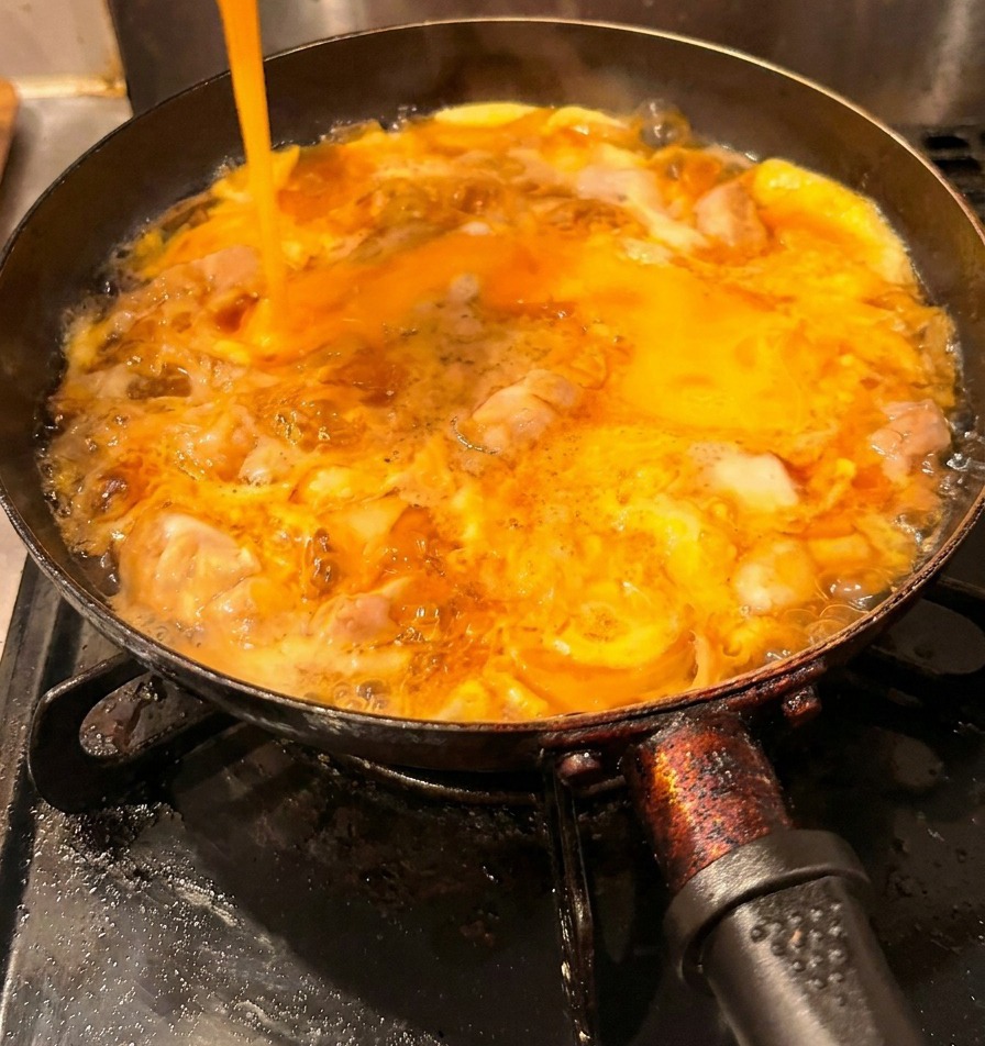 A stream of bright yellow beaten eggs being poured into a pan over chicken and a base layer of cooked eggs.