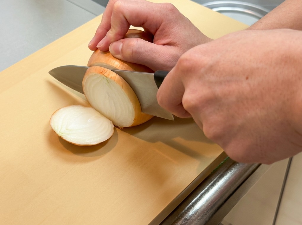 A person cutting a whole peeled yellow onion in half lengthwise on a light-colored cutting board.