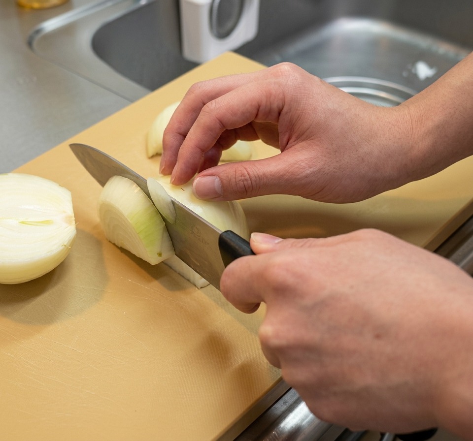Onion halves being sliced into thick longitudinal wedges on a tan cutting board.