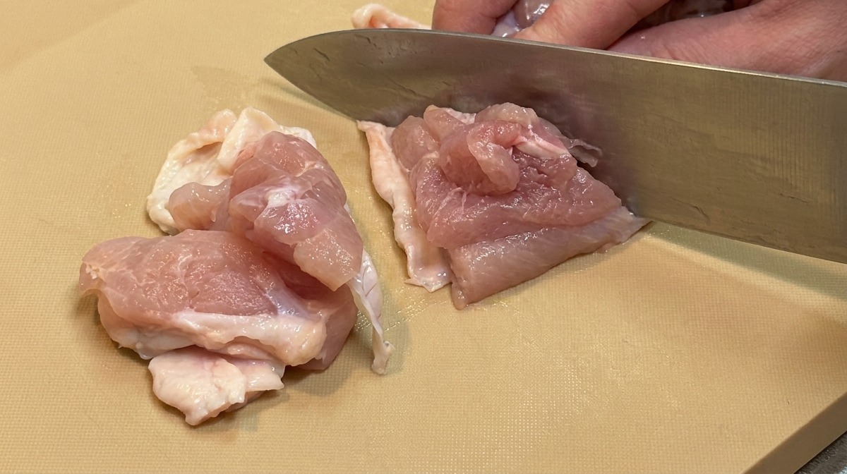 Raw chicken thigh meat being cut into small, even, bite-sized chunks with a large kitchen knife.