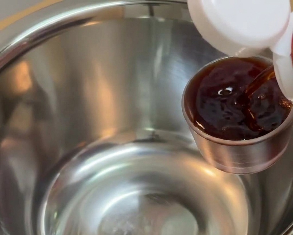 Dark soy sauce being poured from a small metal measuring cup into a larger stainless steel mixing bowl.