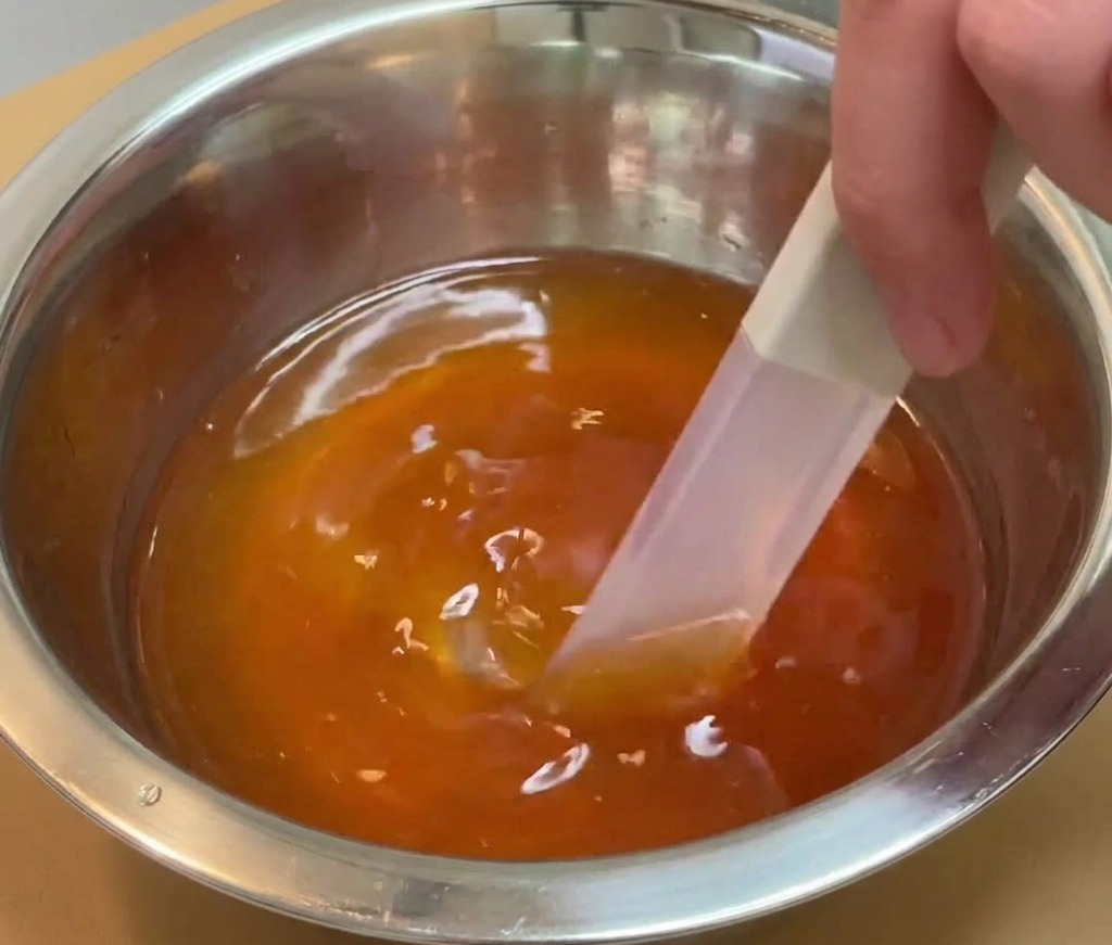 A hand using a spatula to stir a dark, translucent seasoning liquid in a stainless steel mixing bowl.