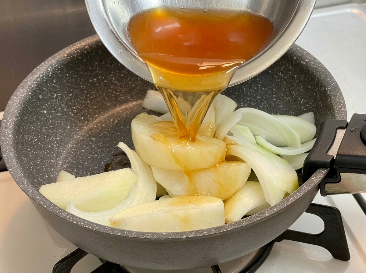 Dark seasoning liquid being poured from a metal bowl over a bed of sliced onions in a frying pan.