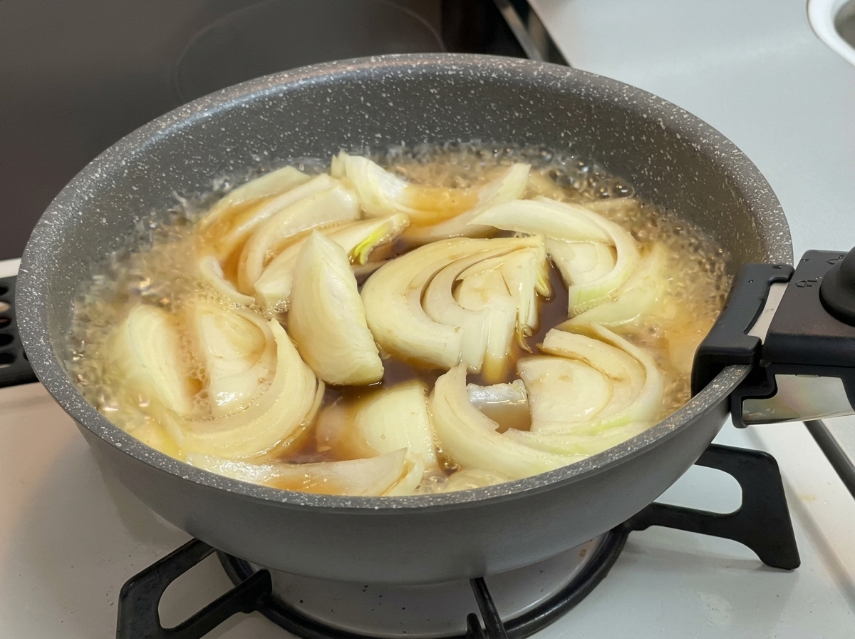 Onions simmering in a bubbling, dark brown broth within a gray frying pan on a stovetop.