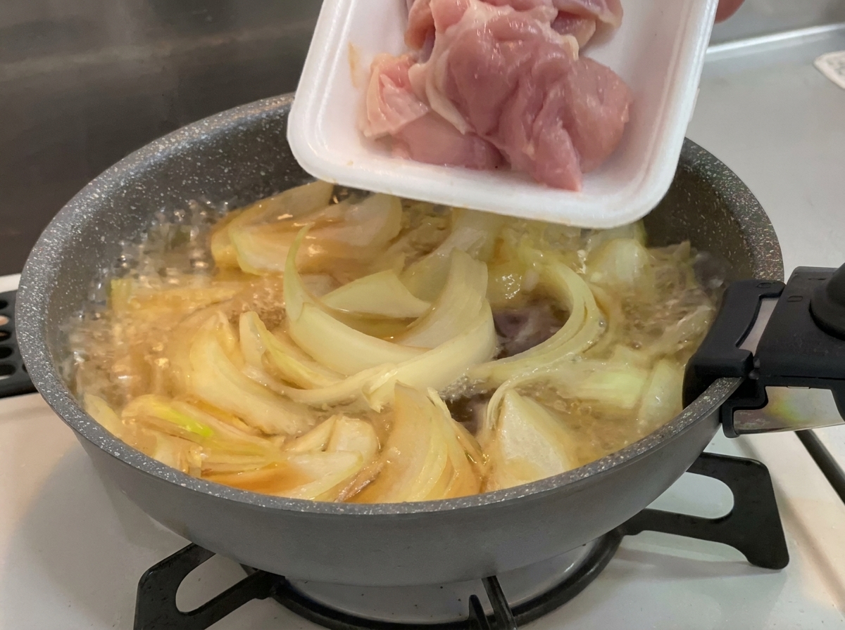 Raw pink chicken thigh pieces being added from a white styrofoam tray into a pan of simmering onions and broth.