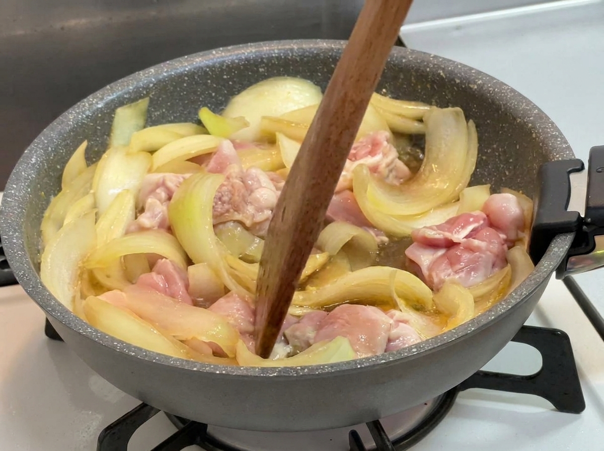 A wooden spatula stirring partially cooked chicken pieces and onions in a simmering dark liquid in a pan.