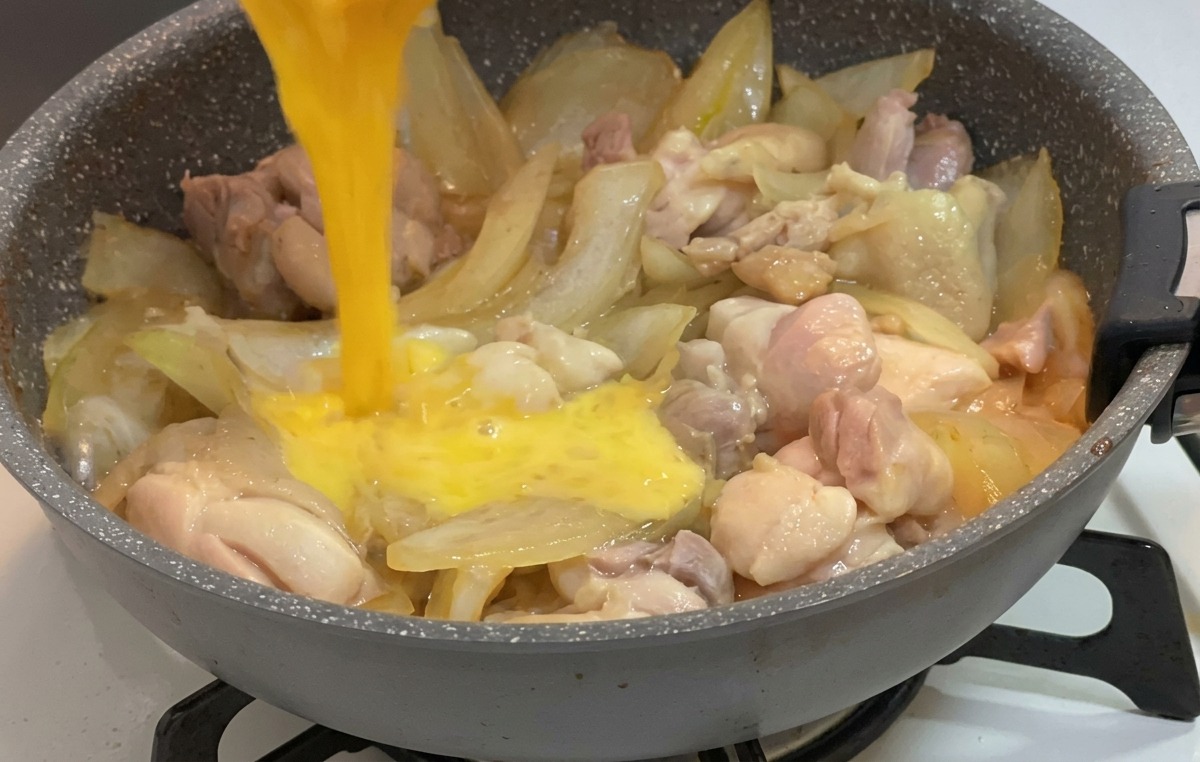 Beaten egg being poured from a small bowl onto chicken and onions simmering in a sauce.
