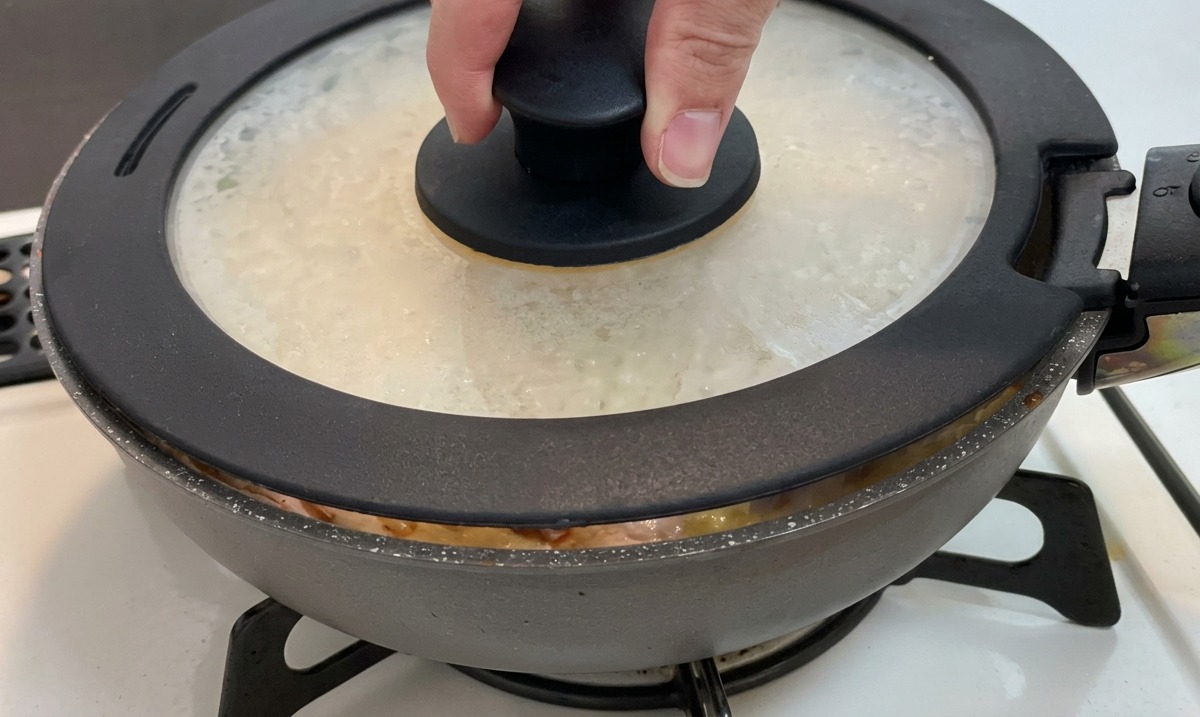 A hand placing a glass lid with a black knob onto a frying pan containing chicken, onions, and eggs.