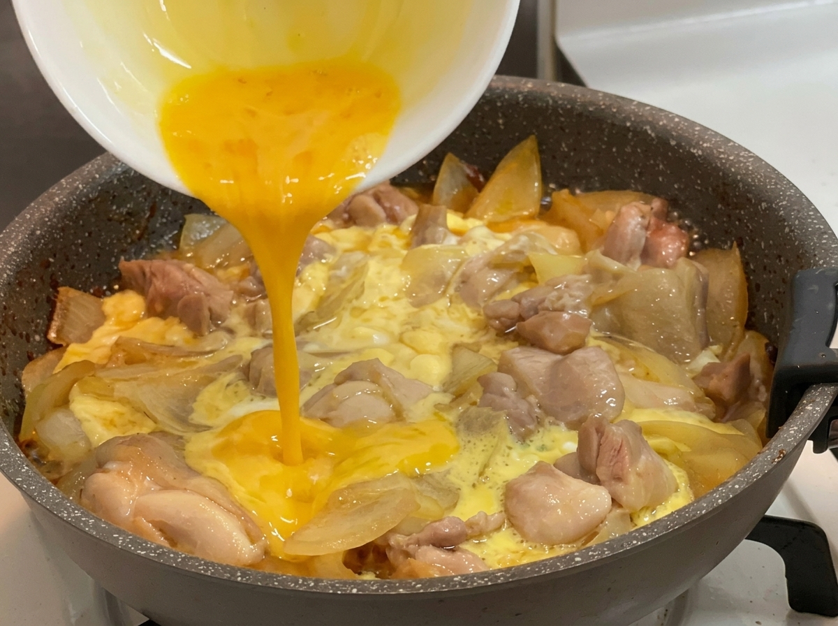 Pouring bright yellow beaten eggs from a white bowl into a hot pan filled with simmering chicken chunks, sliced onions, and partially set eggs.