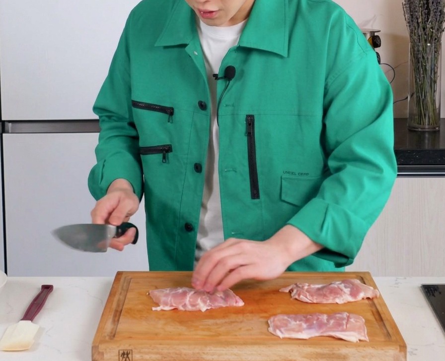 A knife making shallow slits into the meat side of boneless chicken thighs on a wooden cutting board.