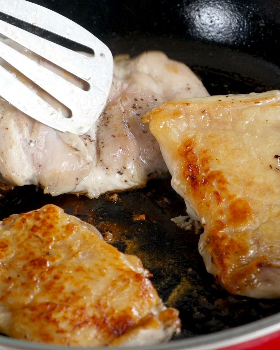 Chicken thighs flipped in a pan, skin-side up showing a golden sear, while the meat side cooks.