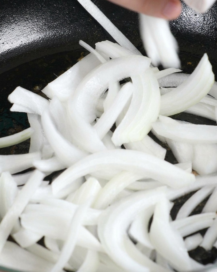 Sliced white onions being dropped into a pan with rendered chicken fat.
