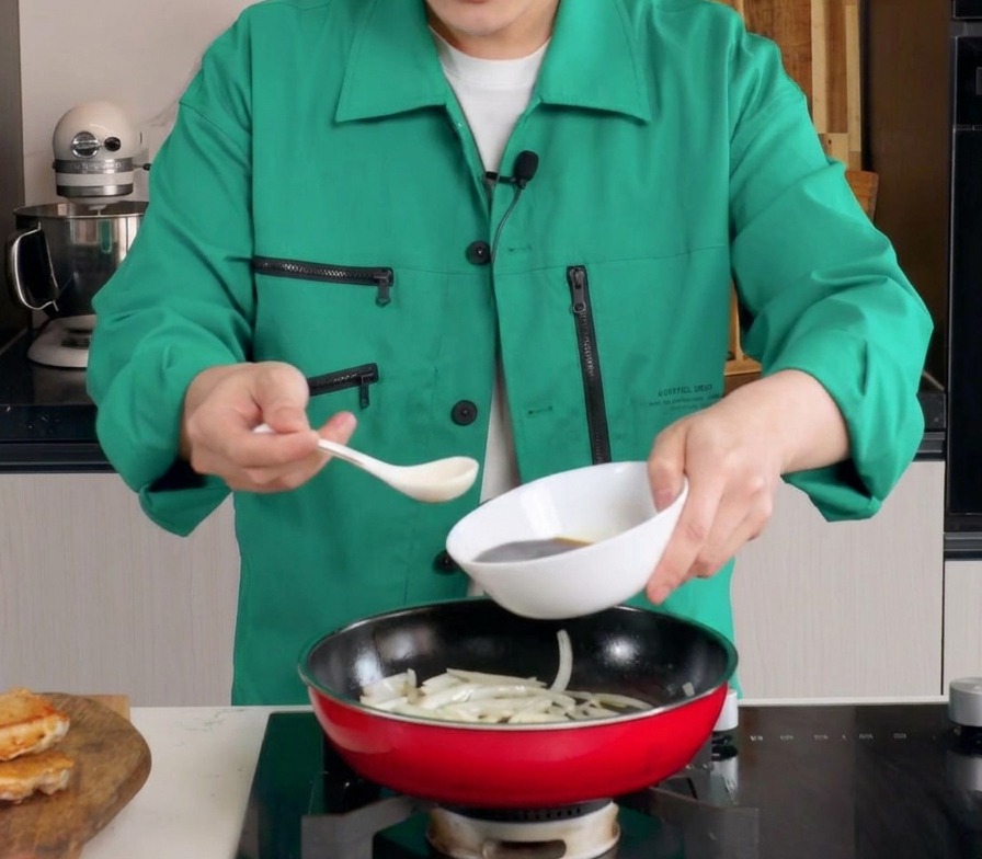 Dark savory sauce being poured from a white bowl into a red frying pan filled with softened onion slices.