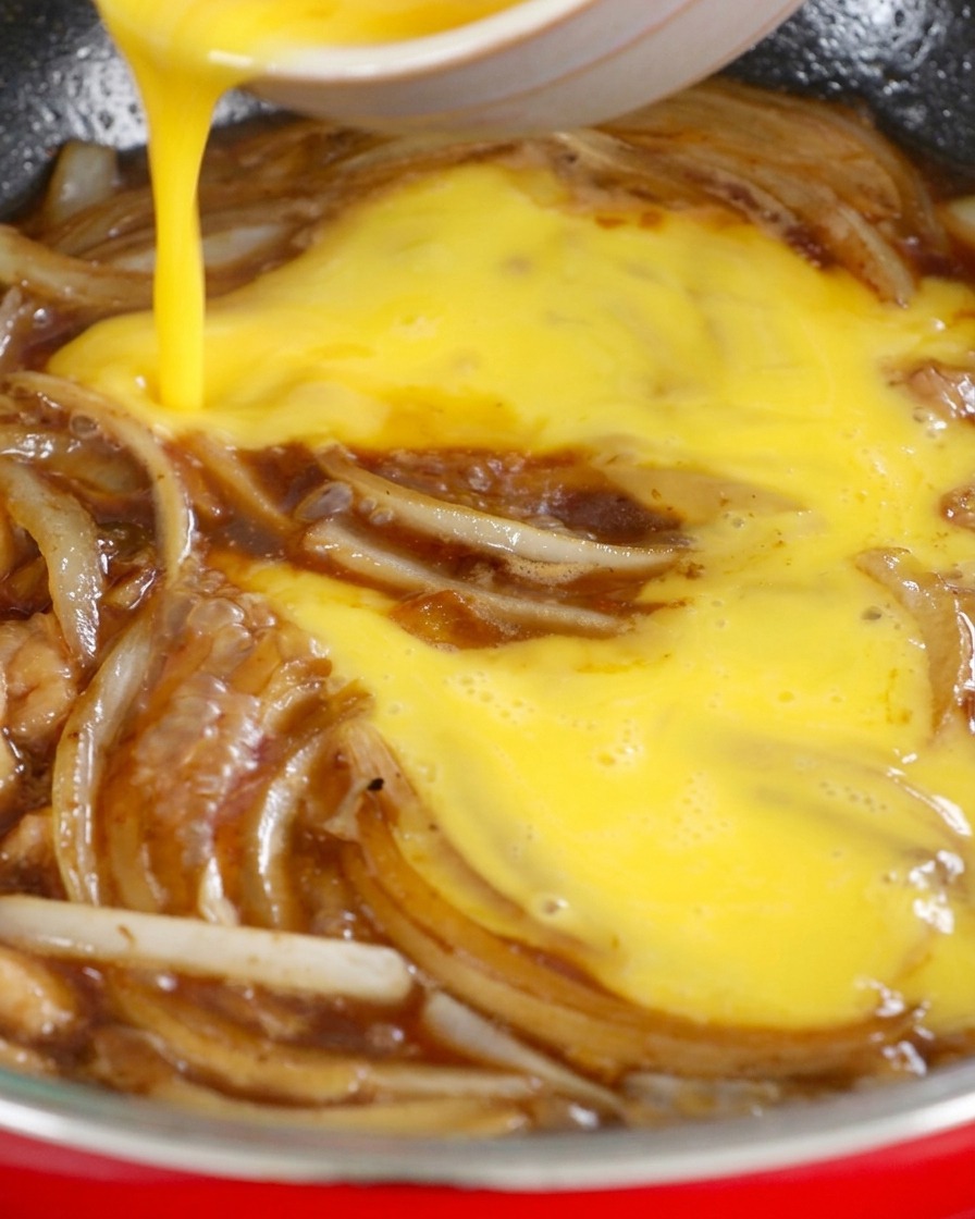 Golden beaten eggs being poured from a bowl into a skillet filled with sliced onions and a dark soy-based sauce.