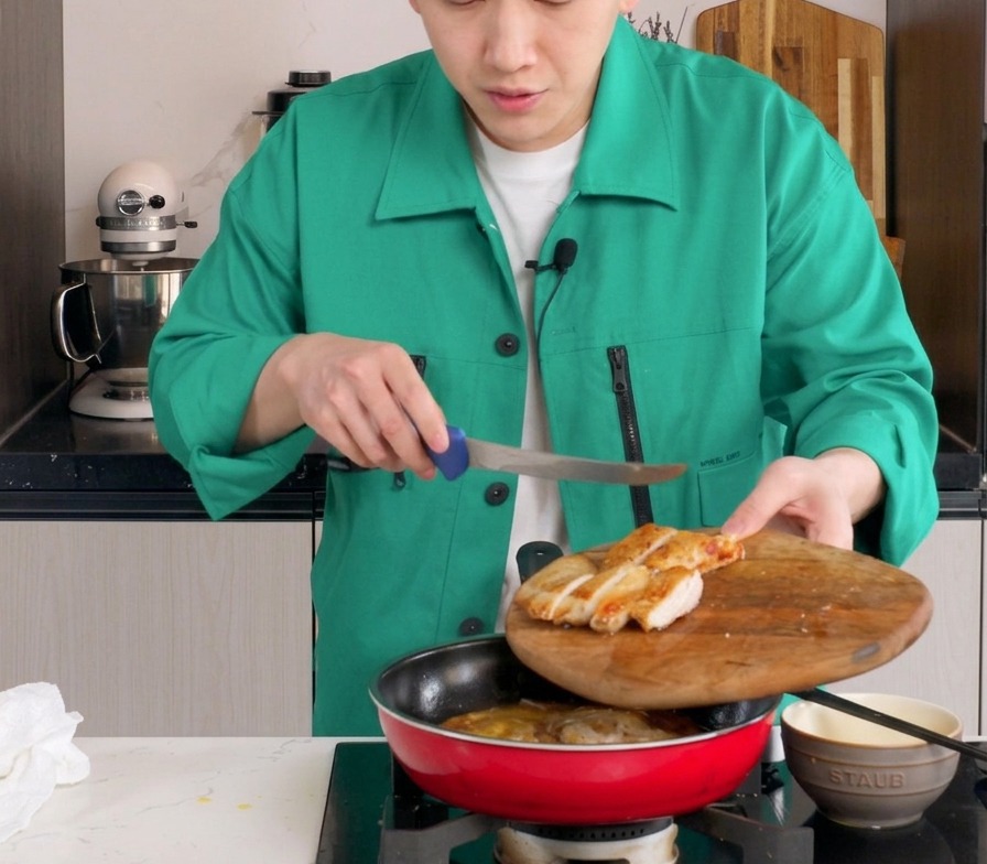 Sliced pan-fried chicken thigh being placed into a red pan containing simmering onions and partially cooked eggs.