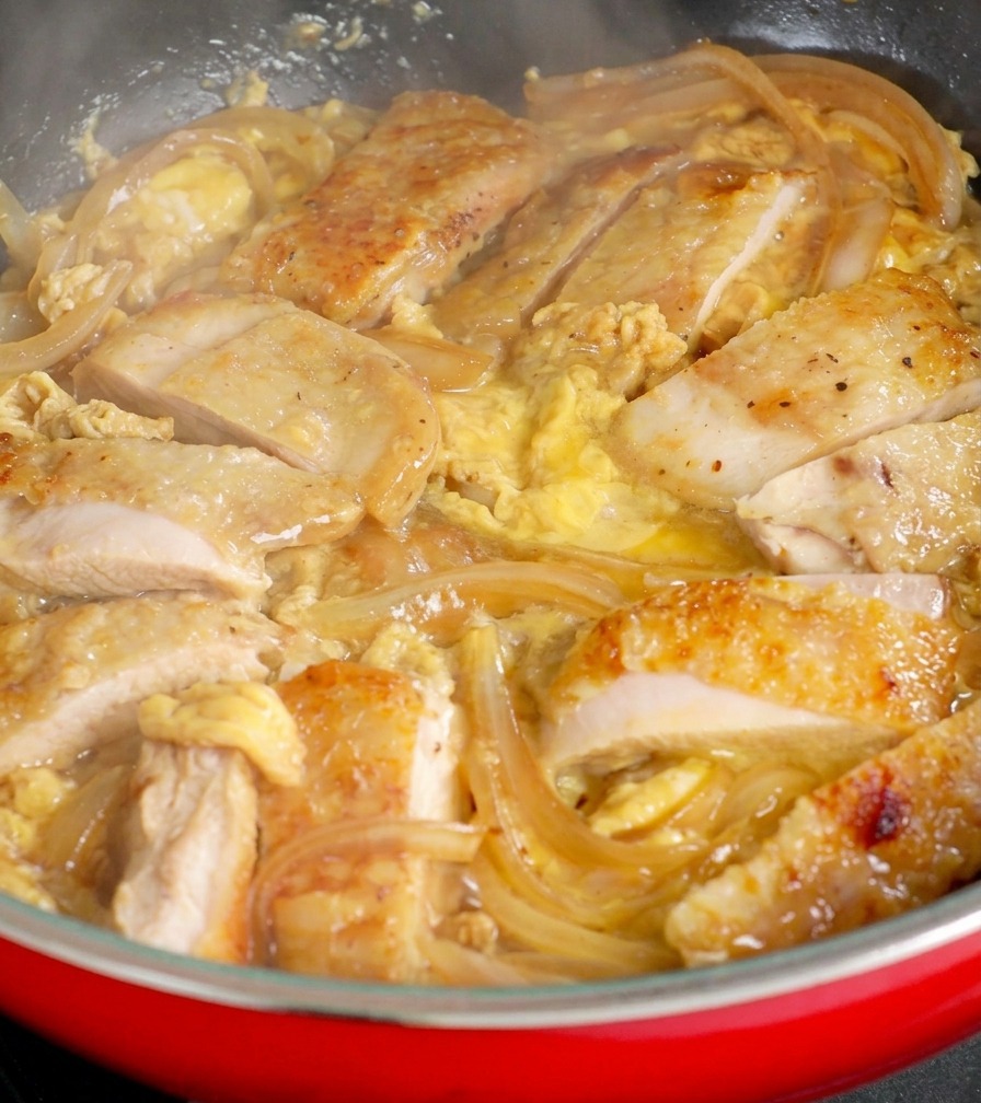 Golden-brown chicken pieces nestled among translucent onions in a red pan, with fresh raw egg being poured over the top to create a custardy sauce.