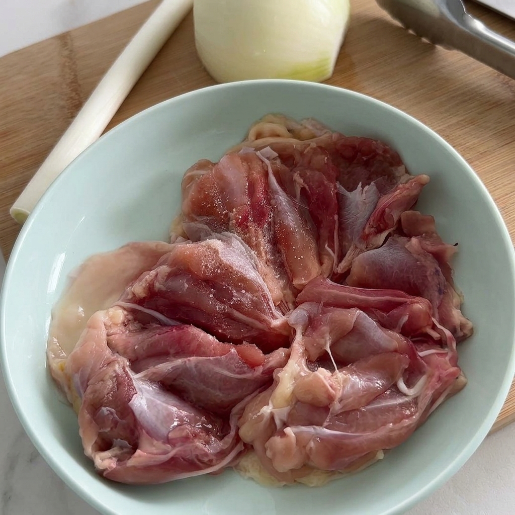 Raw chicken thigh fillets in a pale blue bowl being seasoned with salt and freshly cracked black pepper.