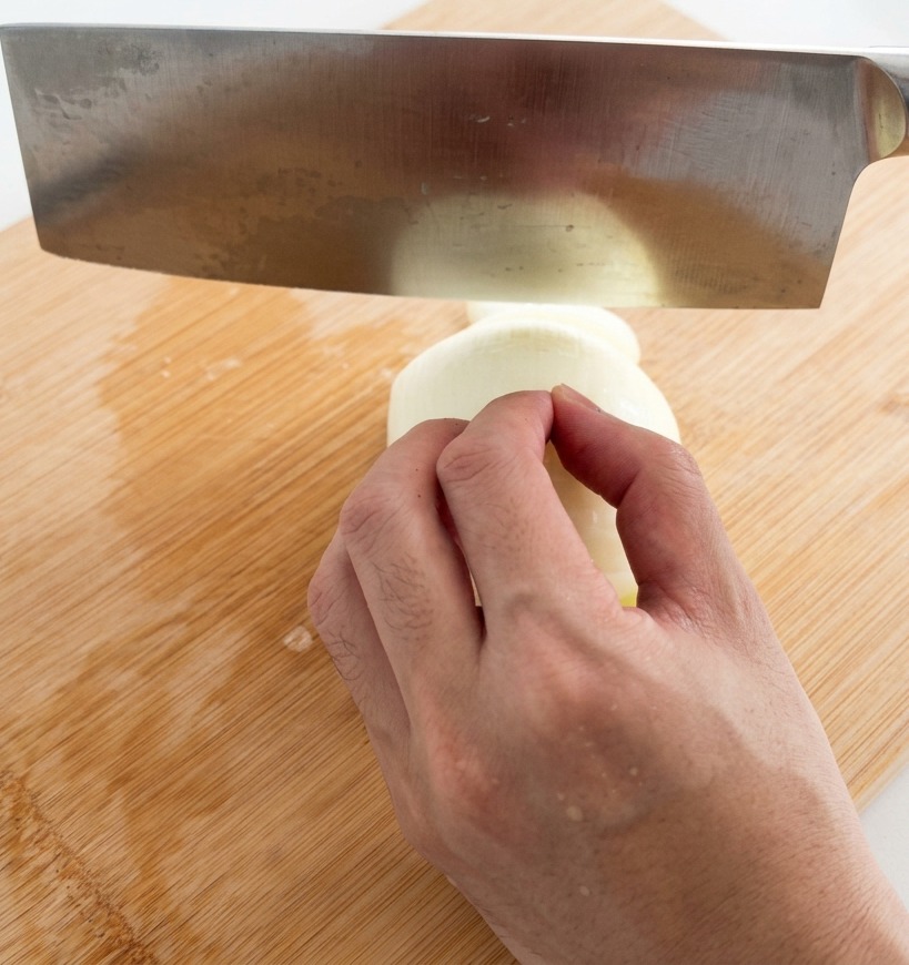 A hand using a large stainless steel cleaver to slice a white onion into thin strips on a bamboo cutting board.