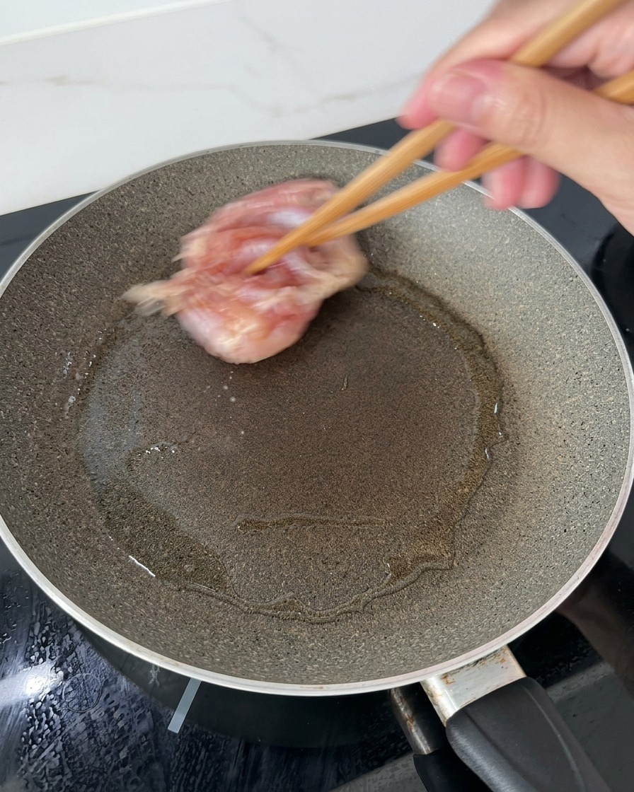 Chicken thighs being lowered into a hot, oiled frying pan using wooden chopsticks, starting with the skin side down.