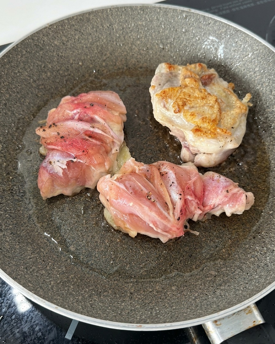 Three chicken thighs in a frying pan, with one flipped over to show a golden-brown, crispy seared surface.